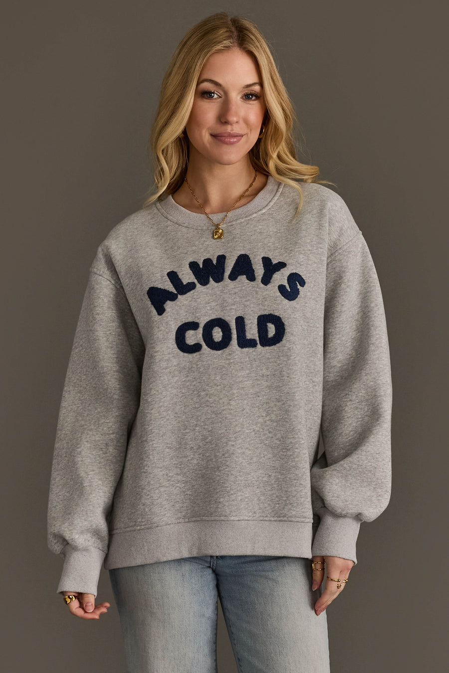 A woman with long blonde hair wears the Panache Apparel Collection’s Always Cold Sweatshirt—a grey pullover featuring “ALWAYS COLD” in large, dark blue letters—while standing against a plain brown background.