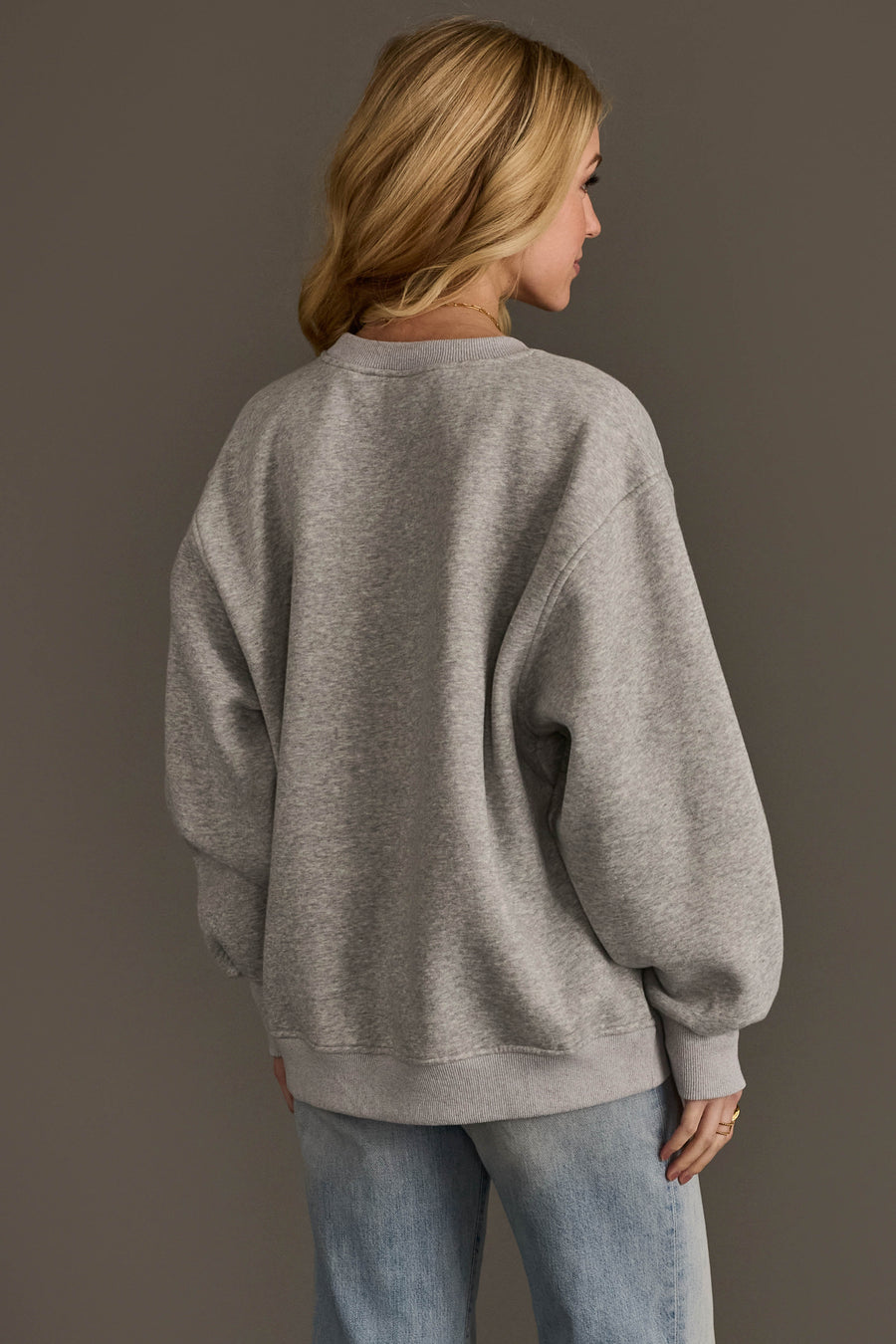 A person with long blonde hair is seen from behind, standing against a plain grey background and wearing the Always Cold Sweatshirt by Panache Apparel Collection in grey, paired with light blue jeans.