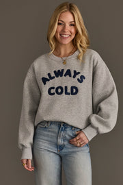A woman smiles while wearing the Panache Apparel Collection’s Always Cold Sweatshirt in gray, featuring “ALWAYS COLD” in navy letters, paired with light blue jeans and standing against a plain dark background.