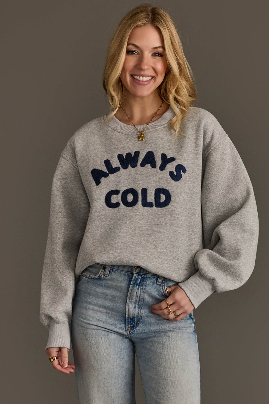 A woman smiles while wearing the Panache Apparel Collection’s Always Cold Sweatshirt in gray, featuring “ALWAYS COLD” in navy letters, paired with light blue jeans and standing against a plain dark background.