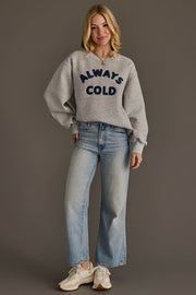 A woman smiles against a plain background, wearing the Panache Apparel Collection’s Always Cold Sweatshirt—grey with “ALWAYS COLD” in navy lettering—paired with light blue wide-leg jeans and white sneakers. Her hands rest relaxed by her sides.