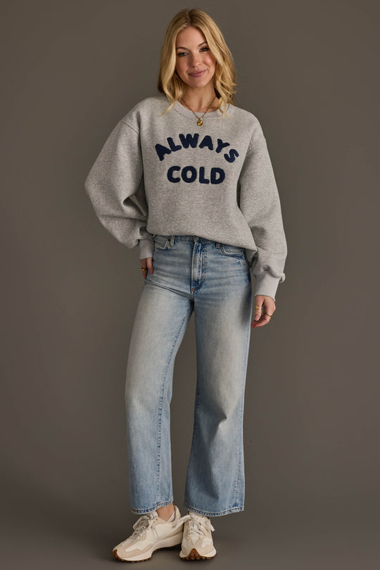 A woman smiles against a plain background, wearing the Panache Apparel Collection’s Always Cold Sweatshirt—grey with “ALWAYS COLD” in navy lettering—paired with light blue wide-leg jeans and white sneakers. Her hands rest relaxed by her sides.