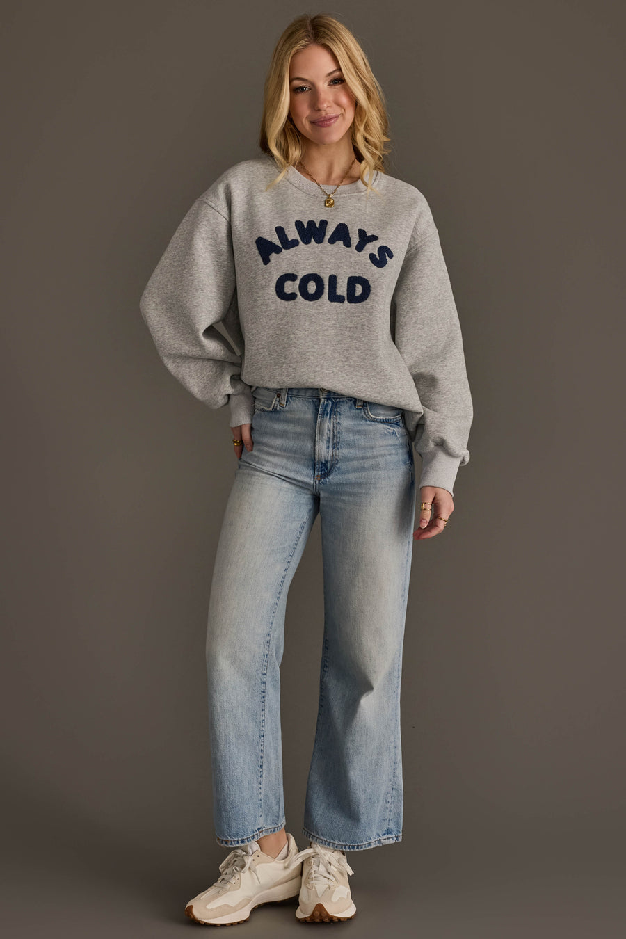 A woman smiles against a plain background, wearing the Panache Apparel Collection’s Always Cold Sweatshirt—grey with “ALWAYS COLD” in navy lettering—paired with light blue wide-leg jeans and white sneakers. Her hands rest relaxed by her sides.
