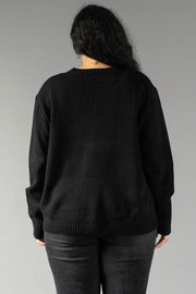 A person with long, dark, wavy hair stands with their back to the camera against a plain gray background. They are wearing the Black Coffee Weather Sweater by Panache Apparel Collection—a long-sleeved, solid black crew-neck sweater—paired with dark gray pants. A thin gold chain necklace is visible around their neck.