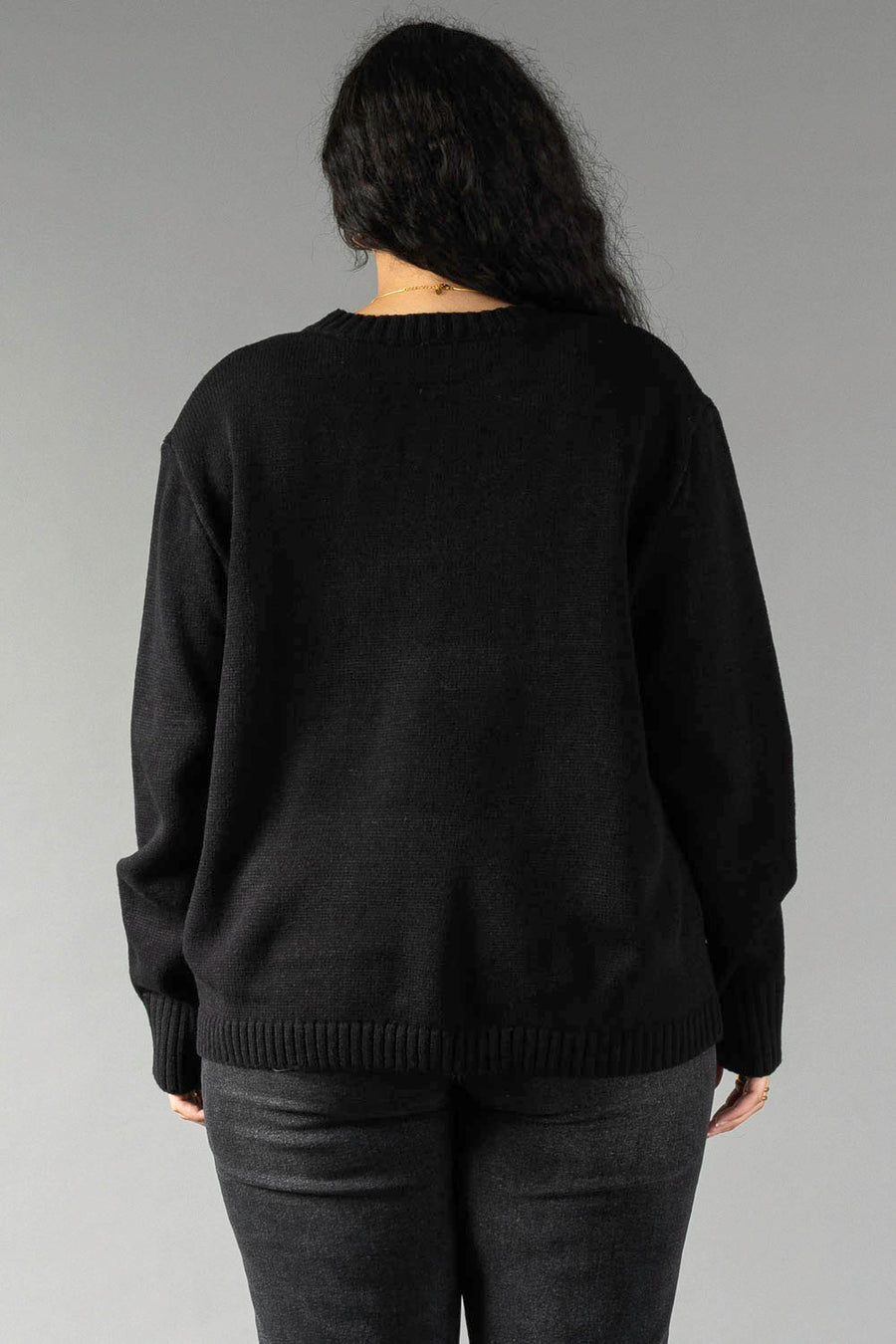 A person with long, dark, wavy hair stands with their back to the camera against a plain gray background. They are wearing the Black Coffee Weather Sweater by Panache Apparel Collection—a long-sleeved, solid black crew-neck sweater—paired with dark gray pants. A thin gold chain necklace is visible around their neck.