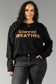 A woman with long curly hair models the Panache Apparel Collection's Black Coffee Weather Sweater, featuring "COFFEE WEATHER" in brown lettering, paired with dark jeans as she poses with one hand in her pocket against a gray background.
