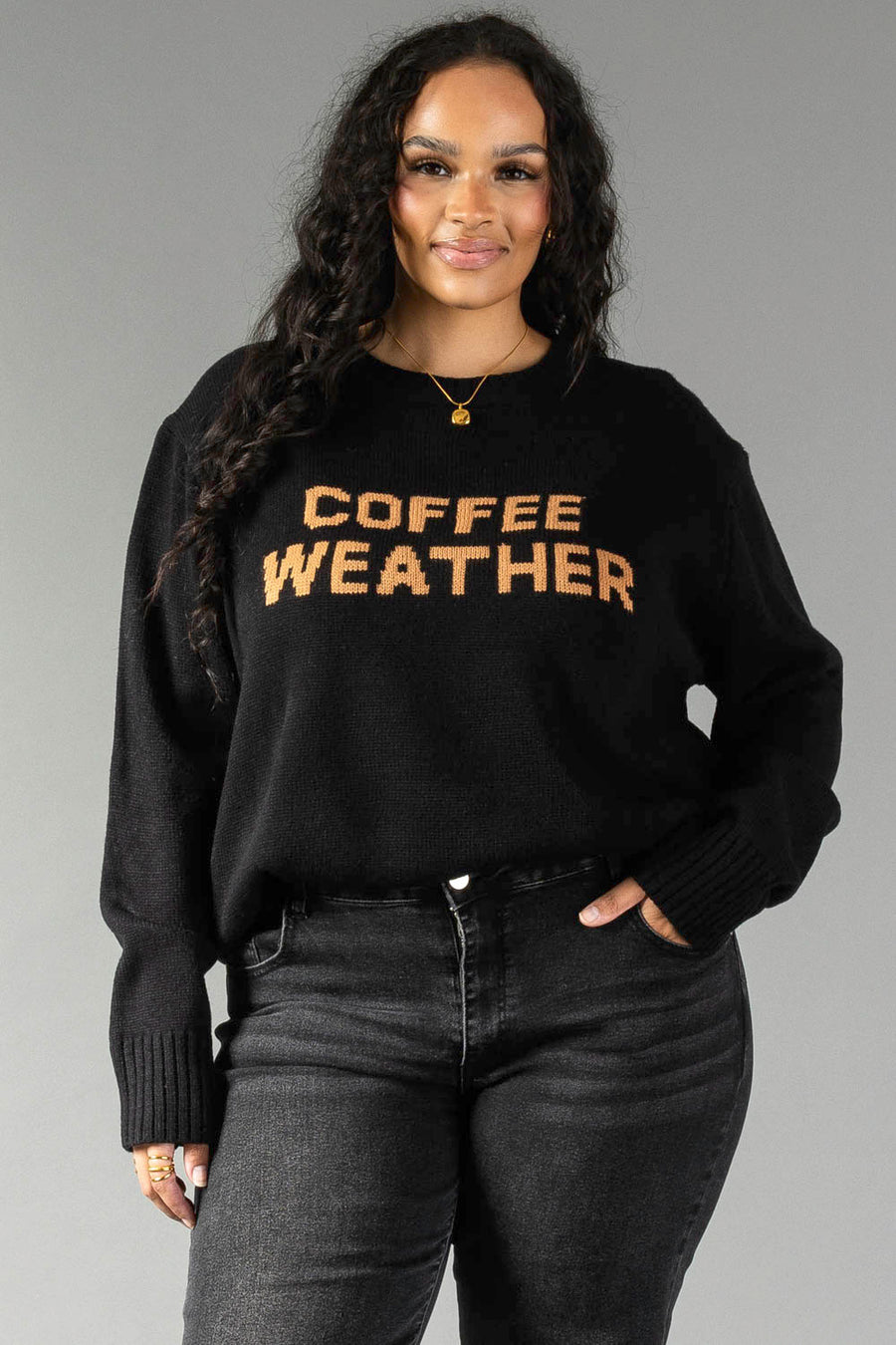 A woman with long curly hair models the Panache Apparel Collection's Black Coffee Weather Sweater, featuring "COFFEE WEATHER" in brown lettering, paired with dark jeans as she poses with one hand in her pocket against a gray background.