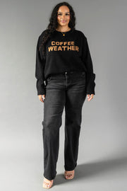 A woman stands against a gray background wearing the Panache Apparel Collection’s Black Coffee Weather Sweater, featuring “COFFEE WEATHER” in orange letters. She pairs it with black jeans and open-toed shoes, her long, wavy hair complementing her confident expression.