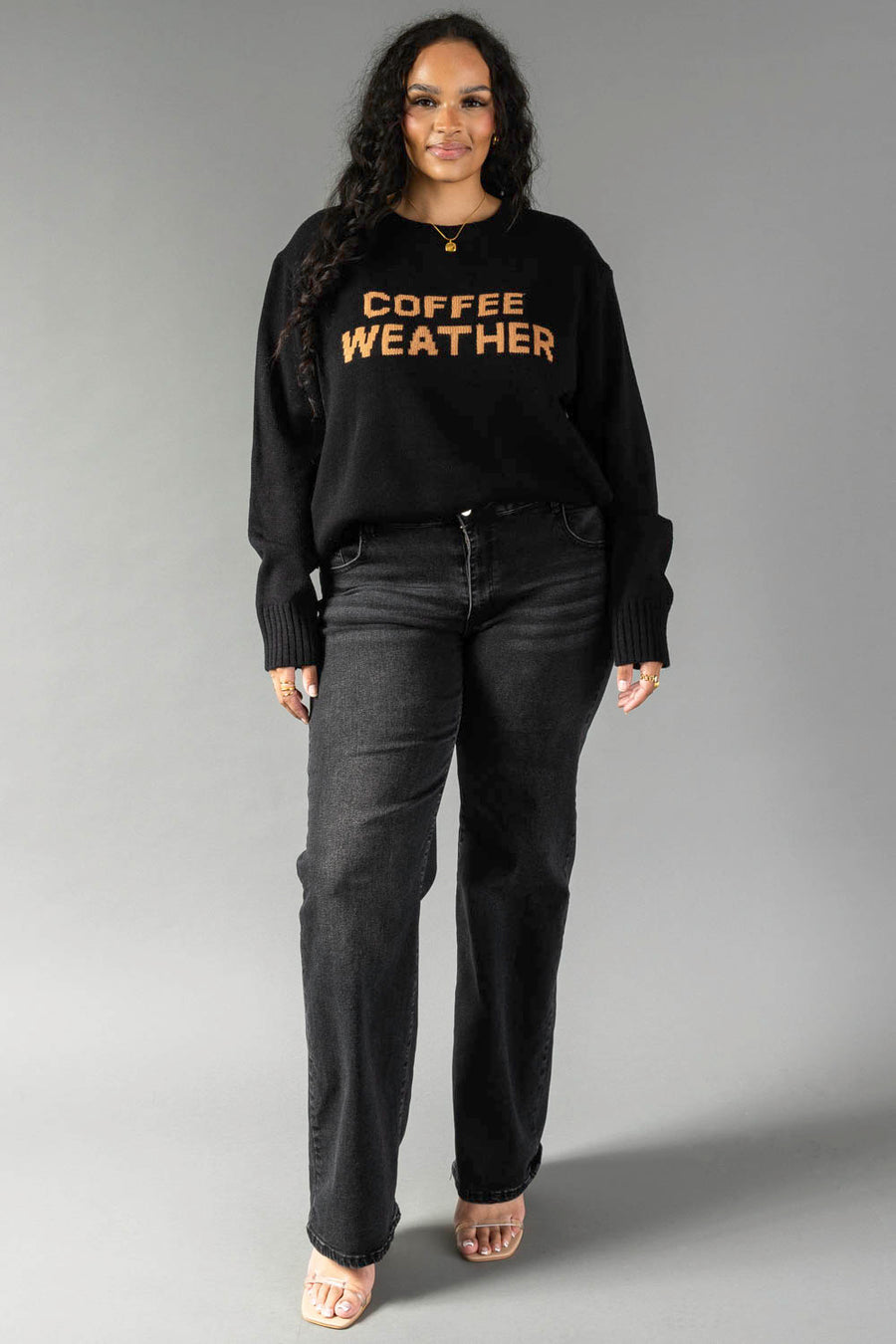 A woman stands against a gray background wearing the Panache Apparel Collection’s Black Coffee Weather Sweater, featuring “COFFEE WEATHER” in orange letters. She pairs it with black jeans and open-toed shoes, her long, wavy hair complementing her confident expression.
