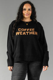 A person with long curly hair wears the Black Coffee Weather Sweater from Panache Apparel Collection, featuring "COFFEE WEATHER" in brown letters, paired with dark pants and standing against a plain gray background.