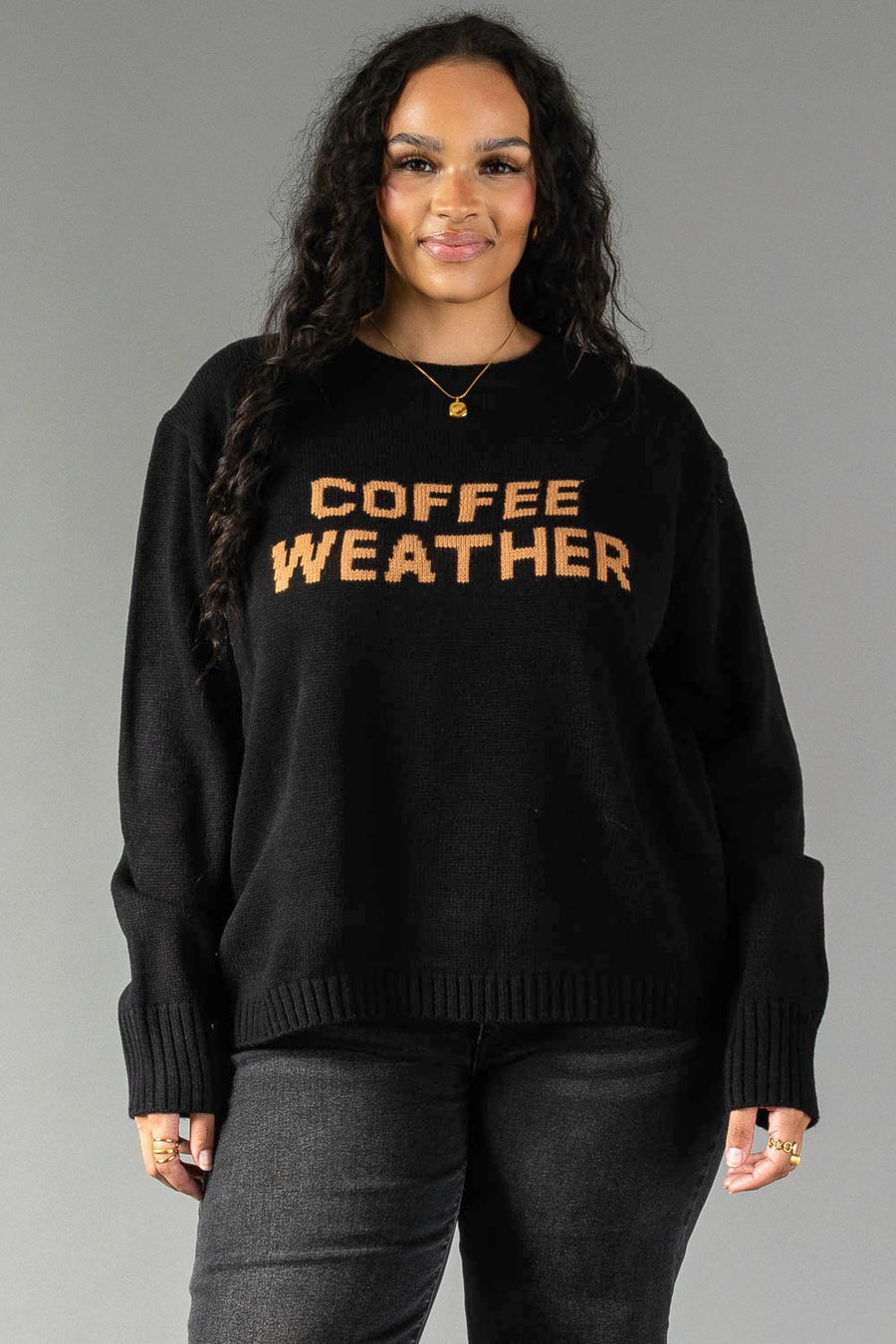 A person with long curly hair wears the Black Coffee Weather Sweater from Panache Apparel Collection, featuring "COFFEE WEATHER" in brown letters, paired with dark pants and standing against a plain gray background.