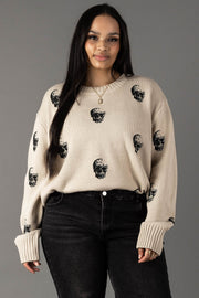 A woman with long dark hair stands against a plain gray background, smiling slightly at the camera while wearing the panacheaccessories Tan Skull Sweater, a relaxed fit, beige knit featuring black skull prints, paired with black jeans and hoop earrings.
