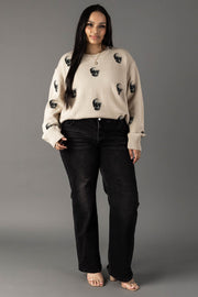 A woman stands against a gray background wearing the Tan Skull Sweater by panacheaccessories, a relaxed fit piece featuring bold black skull designs. She pairs it with black jeans and heeled sandals, her long dark hair framing her relaxed expression as she keeps her hands in her pockets.