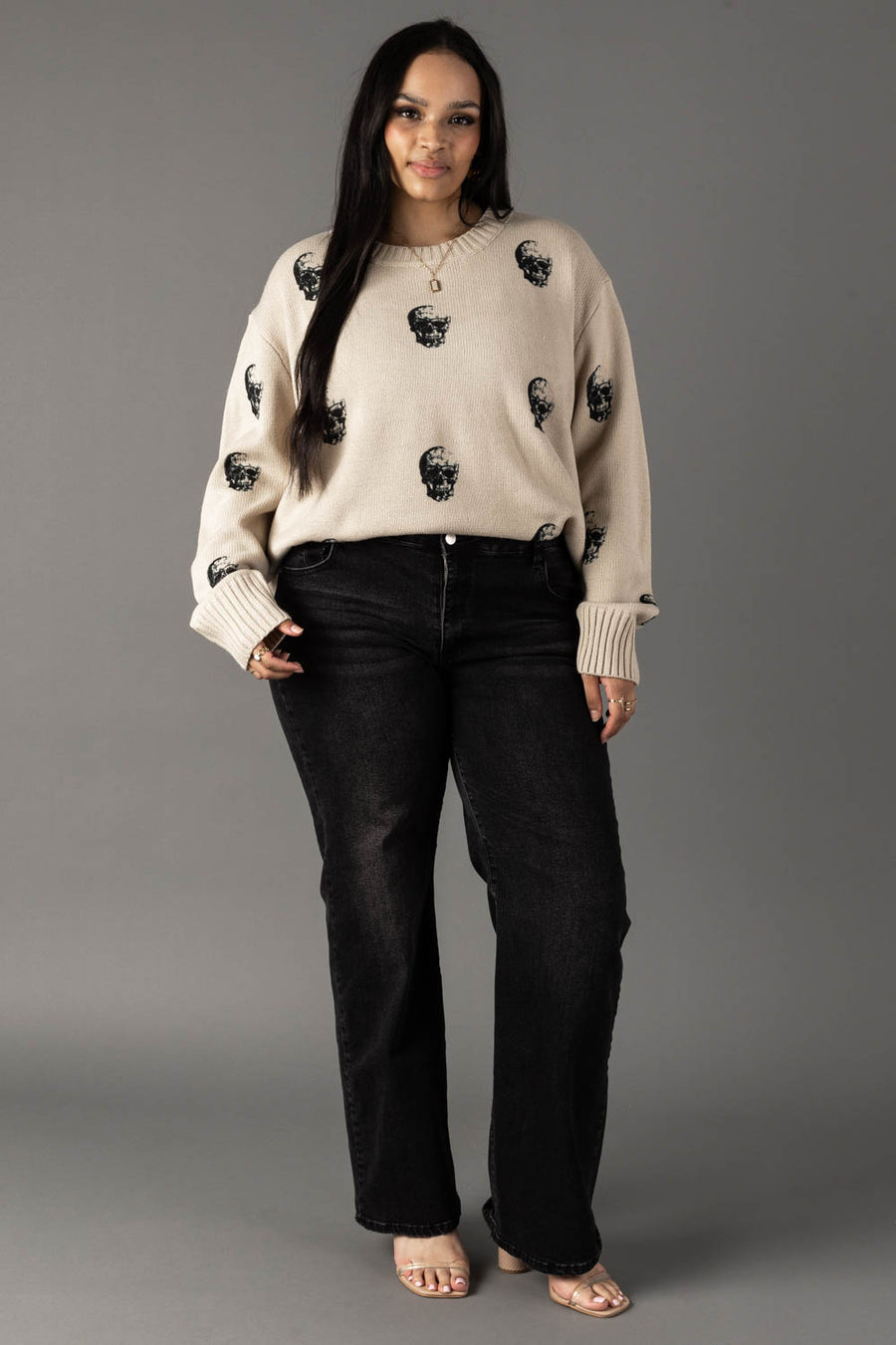 A woman stands against a gray background wearing the Tan Skull Sweater by panacheaccessories, a relaxed fit piece featuring bold black skull designs. She pairs it with black jeans and heeled sandals, her long dark hair framing her relaxed expression as she keeps her hands in her pockets.