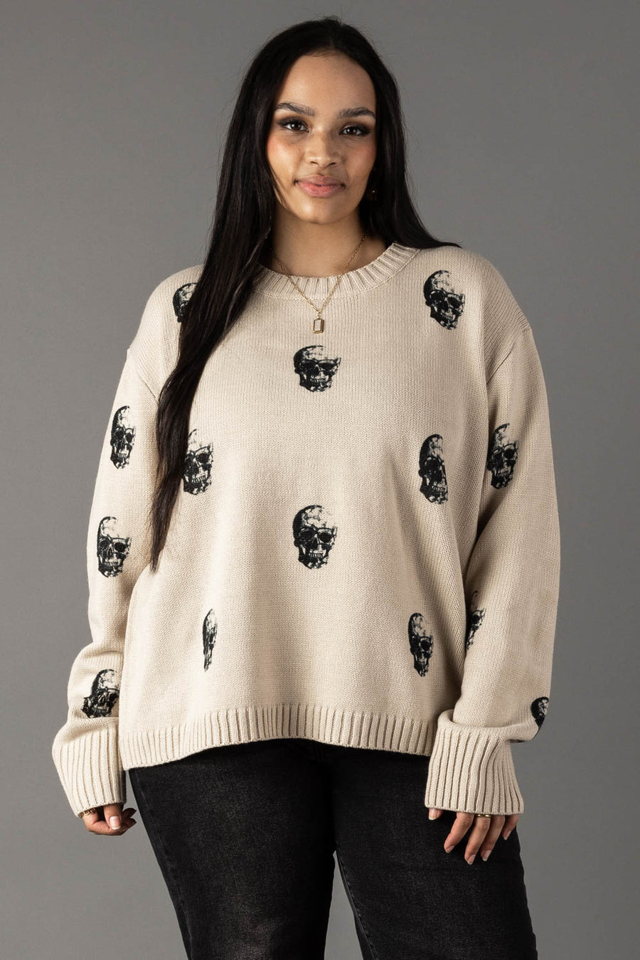 A woman with long dark hair wears the panacheaccessories Tan Skull Sweater with a relaxed fit and black pants, standing against a plain gray background, smiling slightly at the camera.