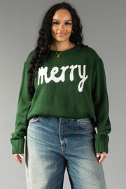 A person wearing a Hunter Green Merry Sweater in hunter green knit with the word merry in white text across the chest, paired with blue jeans.