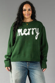 A woman wearing a Hunter Green Merry Sweater, a relaxed fit crewneck in hunter green with the word merry in white script lettering across the chest.