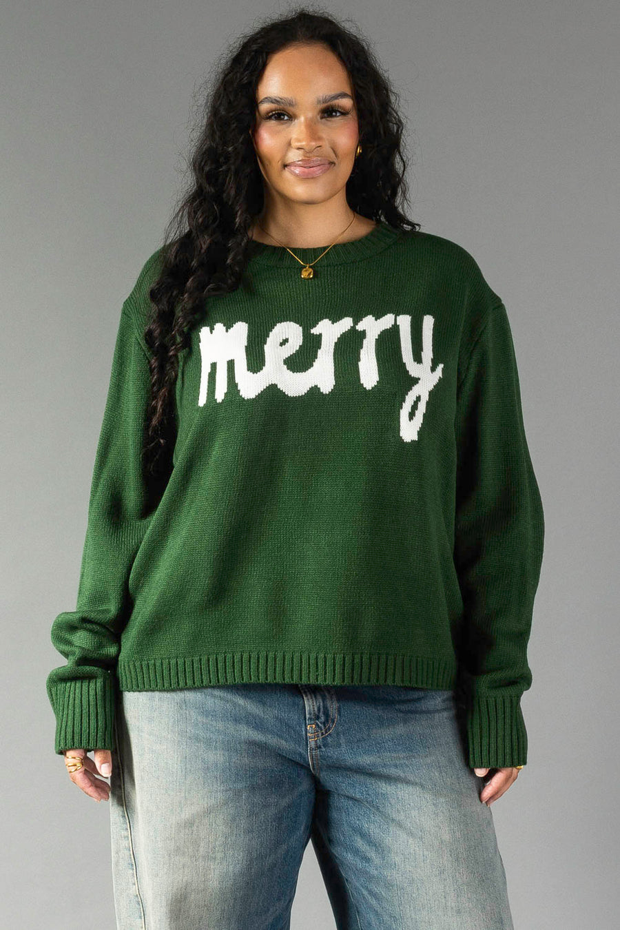 A woman wearing a Hunter Green Merry Sweater, a relaxed fit crewneck in hunter green with the word merry in white script lettering across the chest.