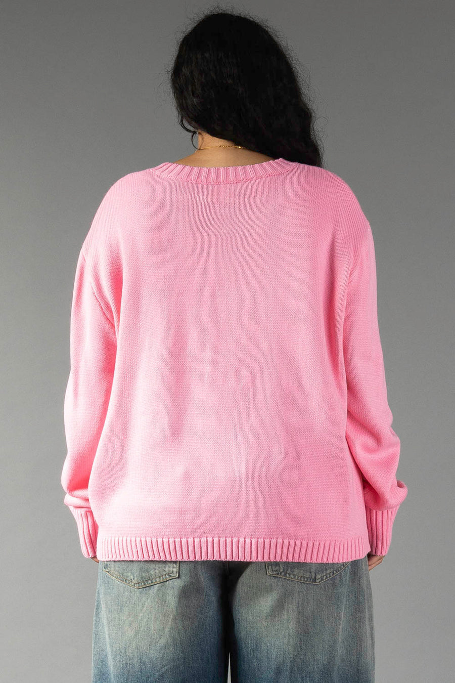 Back view of a person wearing a bright pink relaxed fit Pink & Red Merry Sweater with ribbed trim and blue jeans.
