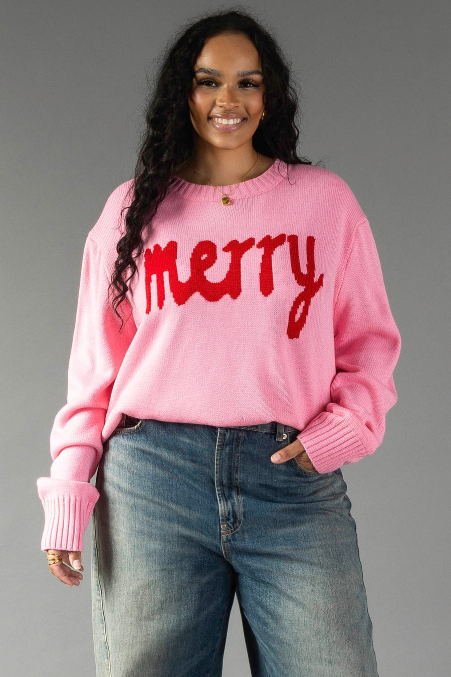 A person wearing a Pink & Red Merry Sweater with the word merry in red text across the chest, paired with blue jeans.