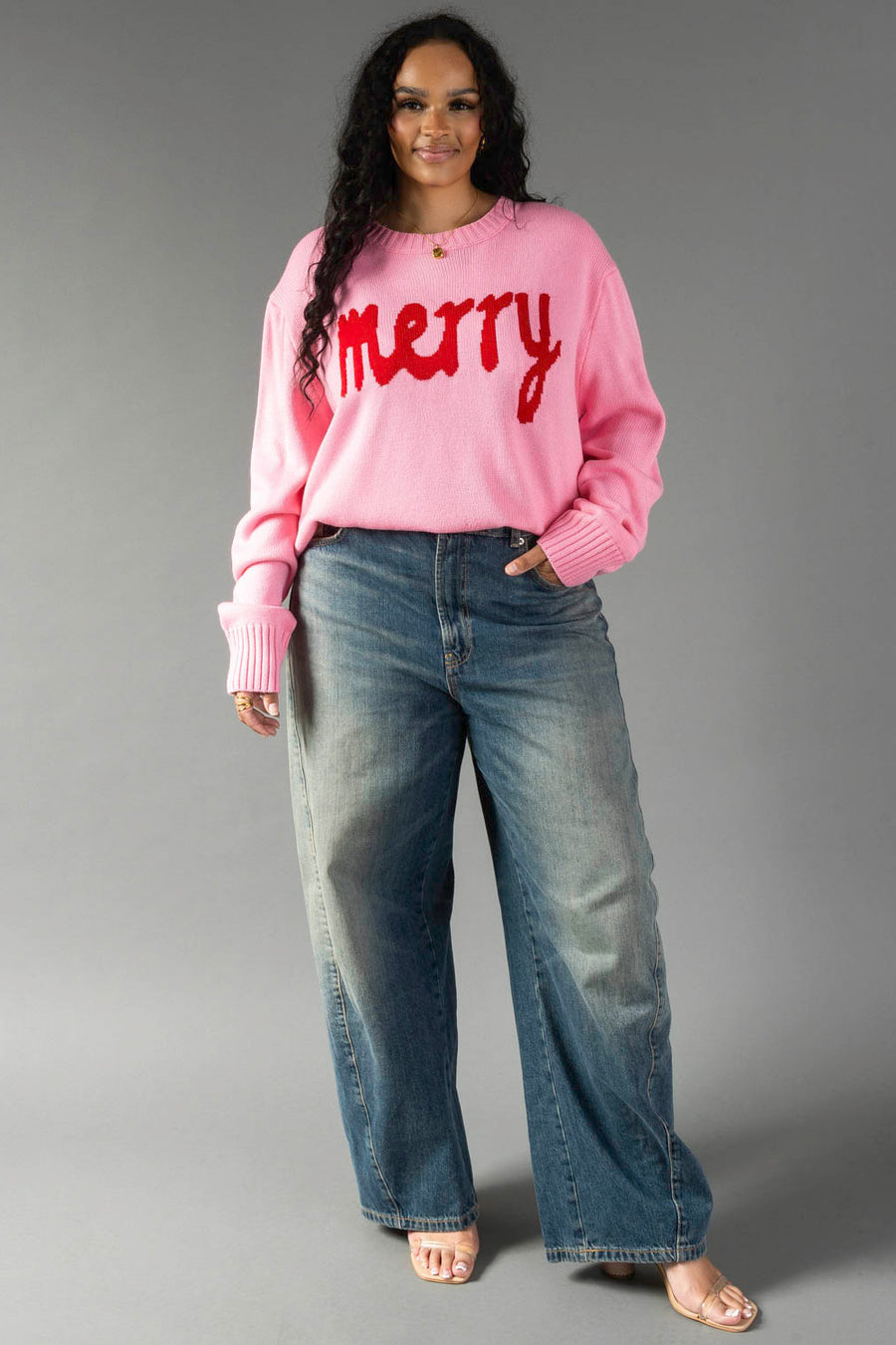 A person wearing a Pink & Red Merry Sweater with red text, paired with wide-leg blue jeans.