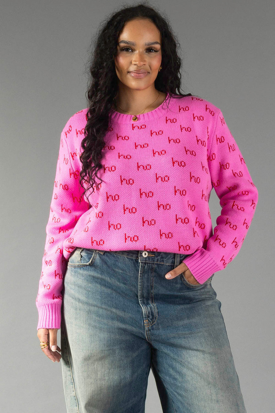 A woman wearing a relaxed fit Pink Ho Ho Ho Sweater with a repeating red print and blue jeans.