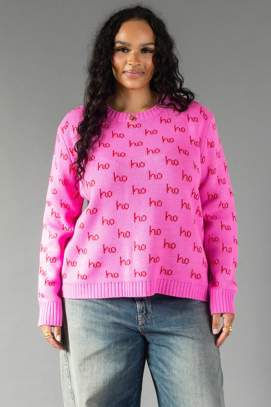 A person wearing a relaxed fit Pink Ho Ho Ho Sweater, featuring a pink knit pattern with the red text ho repeated throughout.