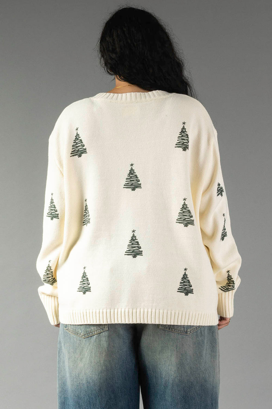 A person wearing an Ivory Festive Tree Sweater, an ivory, relaxed-fit knit sweater patterned with small green Christmas trees.