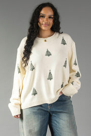 A person wearing a relaxed fit Ivory Festive Tree Sweater in ivory with an all-over pattern of small green Christmas trees and blue jeans.
