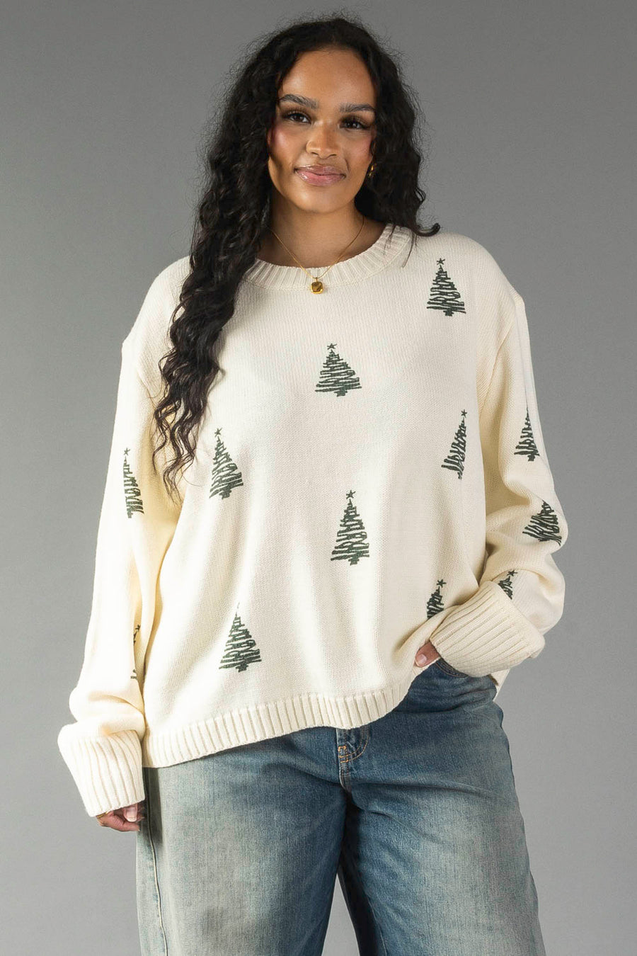 A person wearing a relaxed fit Ivory Festive Tree Sweater in ivory with an all-over pattern of small green Christmas trees and blue jeans.