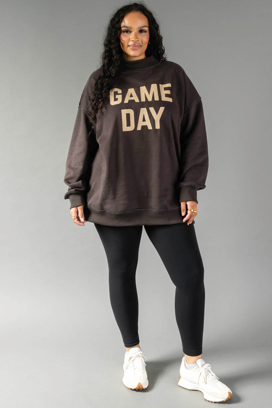 A woman stands against a gray background in the Panache Apparel Collection Dark Gray Game Day & Sunday Funday Reversible Sweatshirt, styled with black leggings and white sneakers.
