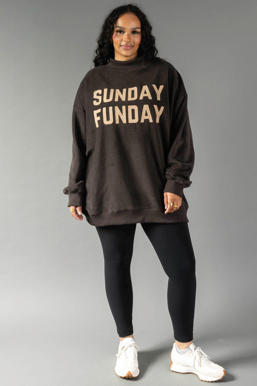 Woman wearing a Dark Gray Game Day & Sunday Funday Reversible Sweatshirt by Panache Apparel Collection, black leggings, and white sneakers, smiling against a gray background.