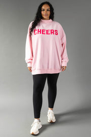 A person wearing a Pink Merry & Cheers Reversible Sweatshirt with the word CHEERS in dark pink letters, black leggings, and white sneakers.