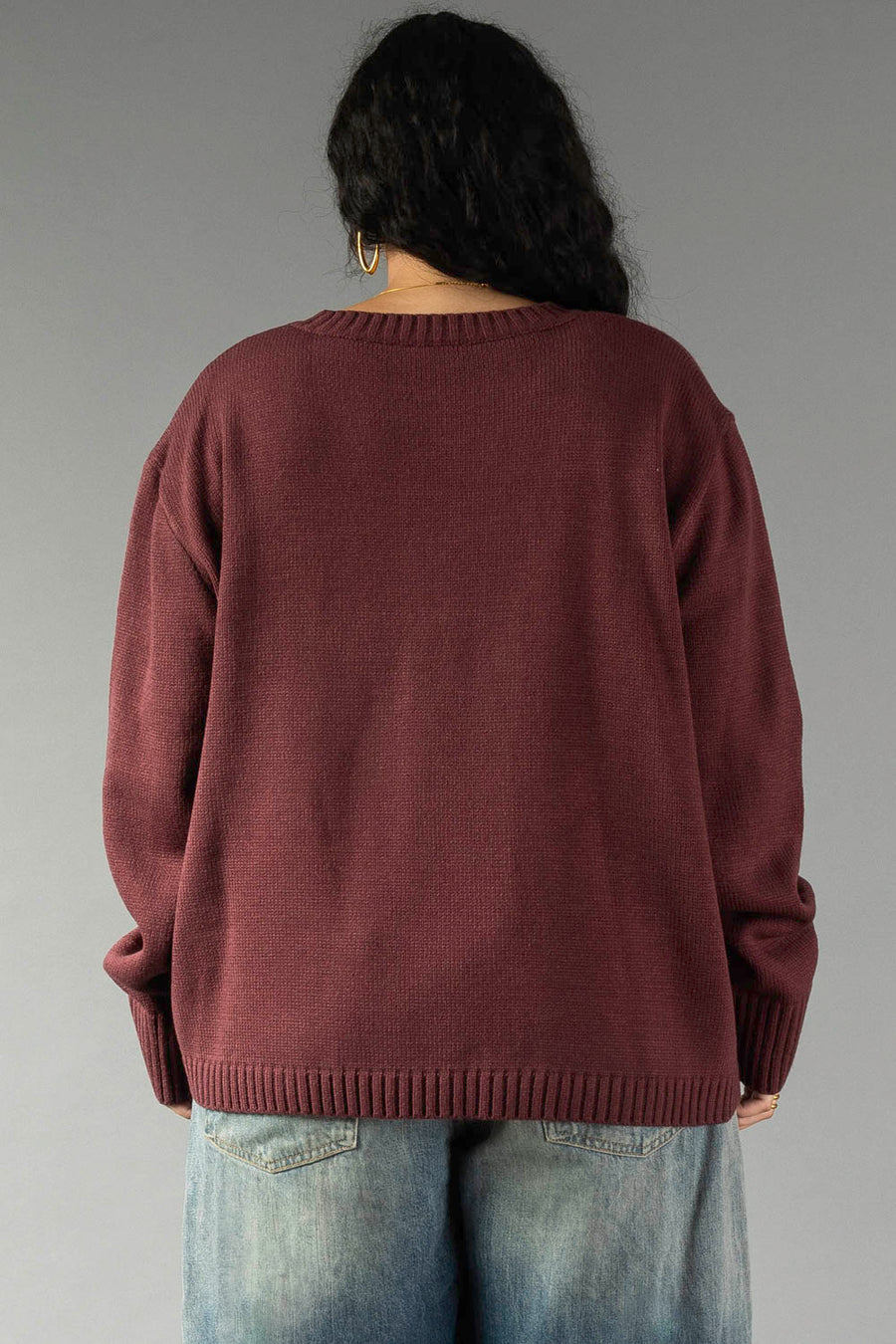 A person with long dark hair is shown from behind, wearing the Panache Apparel Collection Burgundy & Light Pink Wine Season Sweater paired with blue jeans, standing against a plain gray backdrop.
