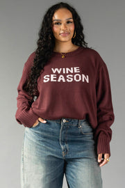 A woman with long curly hair wears the Panache Apparel Collection Burgundy & Light Pink Wine Season Sweater with blue jeans. She is smiling slightly and stands against a plain gray background.