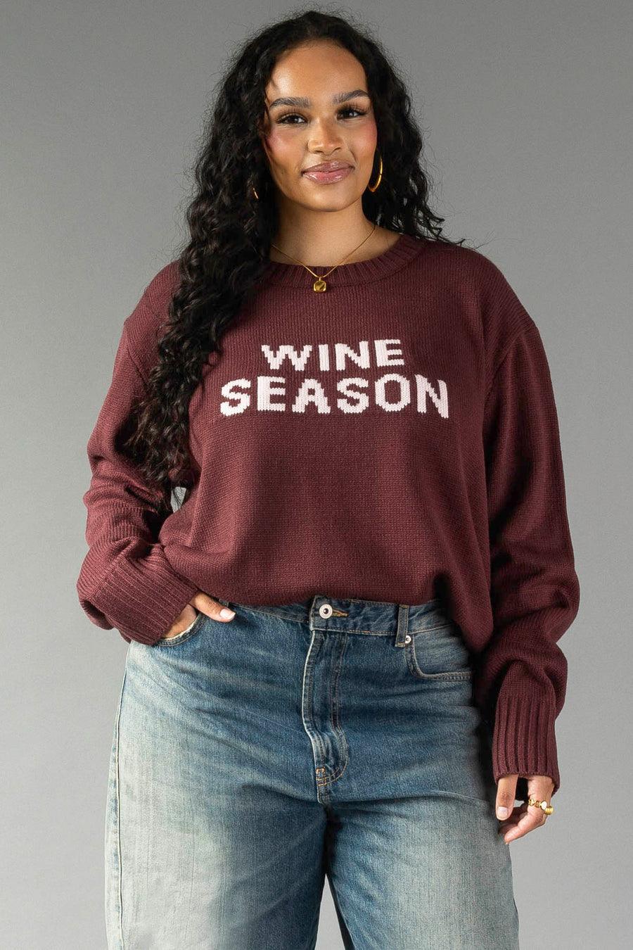 A woman with long curly hair wears the Panache Apparel Collection Burgundy & Light Pink Wine Season Sweater with blue jeans. She is smiling slightly and stands against a plain gray background.