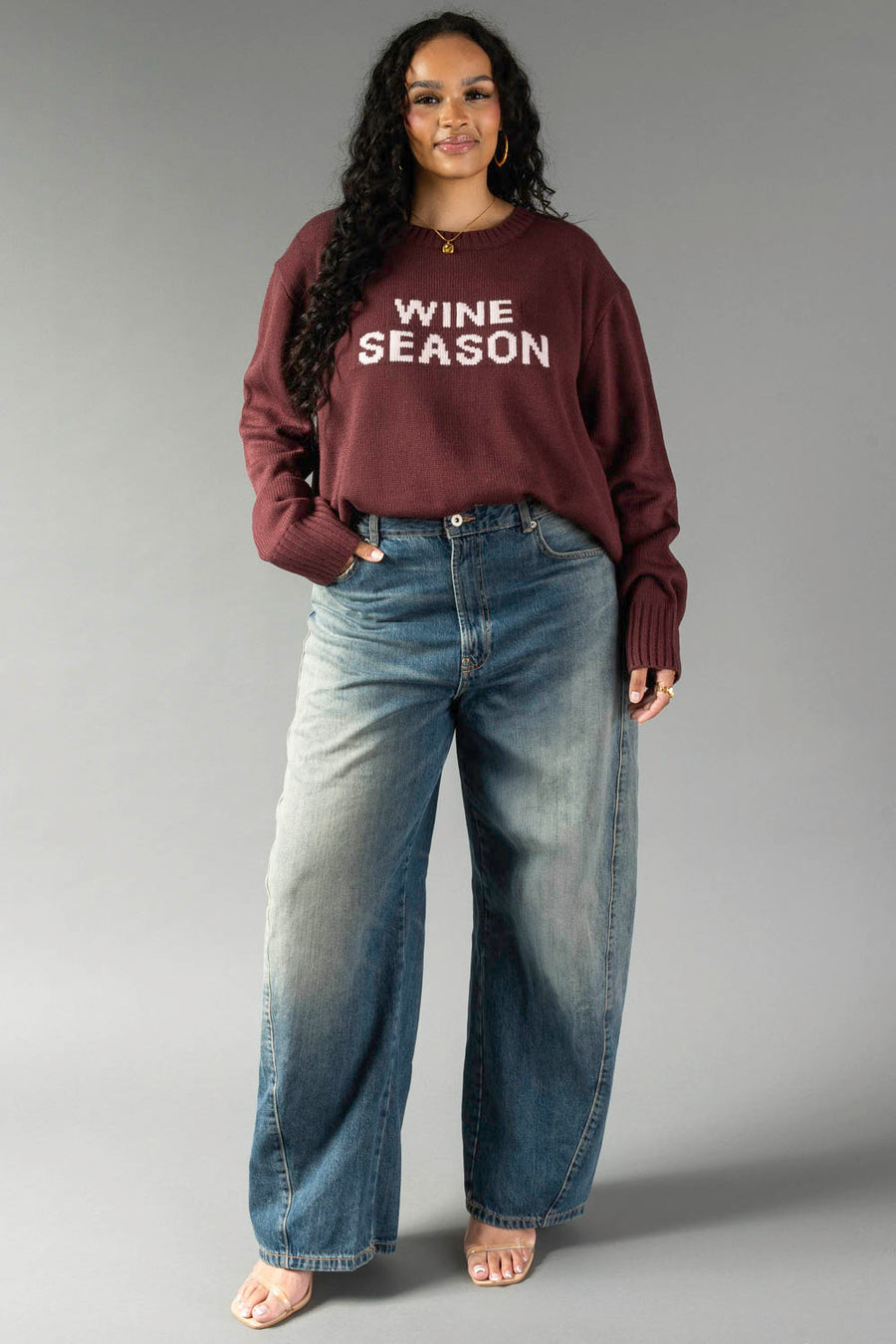 A woman stands confidently against a plain gray background, smiling with one hand in her pocket. She is wearing the Burgundy & Light Pink Wine Season Sweater from Panache Apparel Collection and wide-leg jeans.
