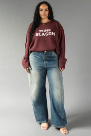 A woman stands against a gray background, wearing the Panache Apparel Collection's Burgundy & Light Pink Wine Season Sweater, paired with blue wide-leg jeans, hoop earrings, and open-toe shoes.