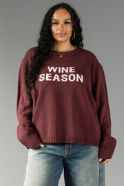 A woman with long curly hair is wearing the Panache Apparel Collection Burgundy & Light Pink Wine Season Sweater, paired with blue jeans, as she stands against a plain gray background.