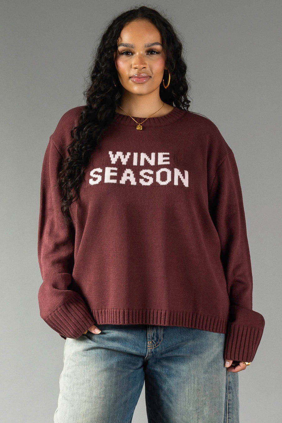 A woman with long curly hair is wearing the Panache Apparel Collection Burgundy & Light Pink Wine Season Sweater, paired with blue jeans, as she stands against a plain gray background.