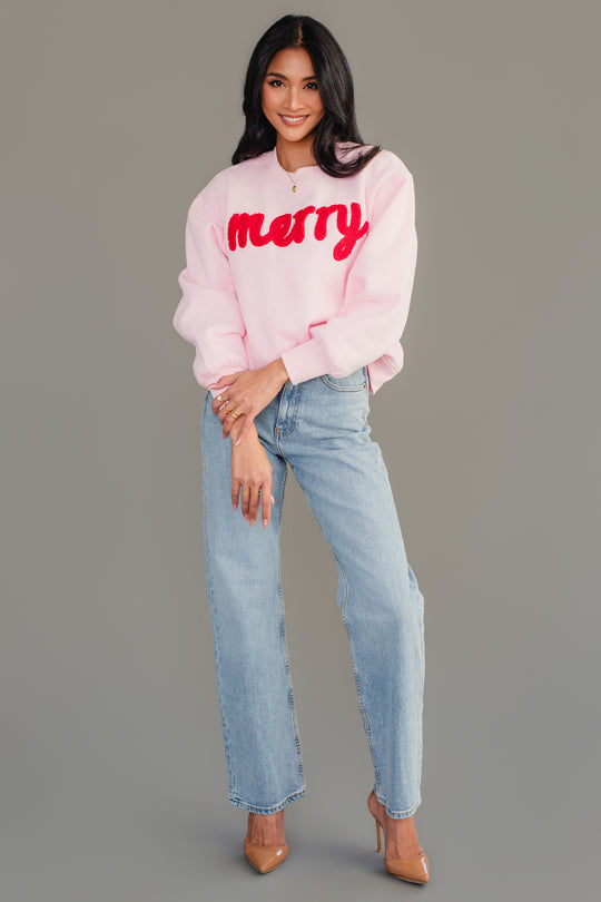 A person wears a Light Pink & Red Merry Sweatshirt with light-wash blue jeans and tan high-heeled pumps.