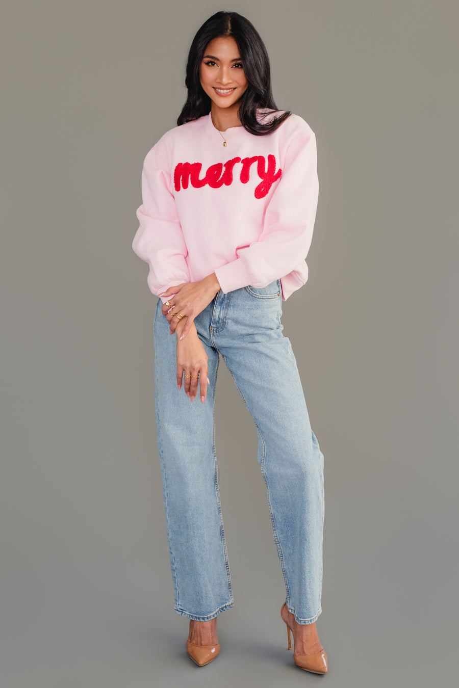 A person wears a Light Pink & Red Merry Sweatshirt with light-wash blue jeans and tan high-heeled pumps.