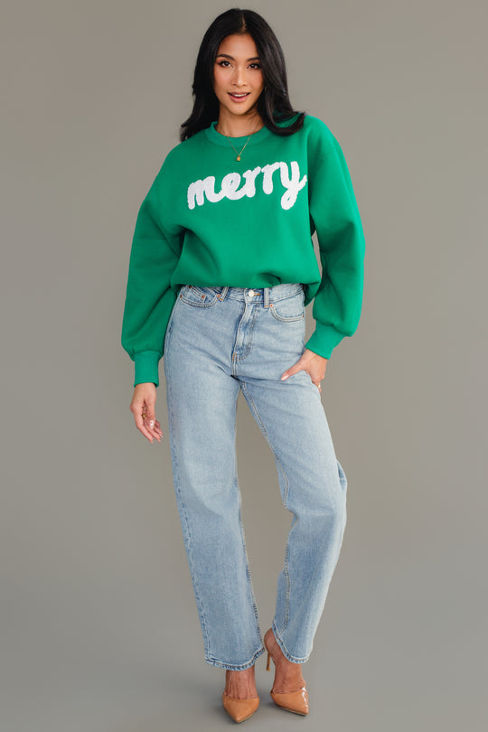 A woman wearing a Green & White Merry Sweatshirt with light-wash blue jeans and tan heels.