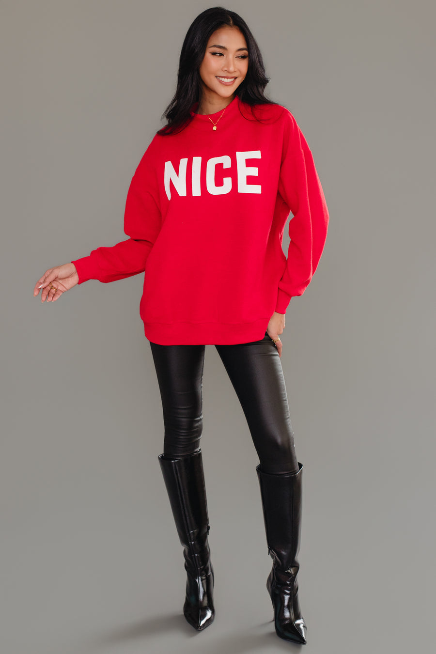 A woman wearing a red Panache Apparel Collection Red Nice & Naughty Reversible Sweatshirt with the word NICE printed in white, black pants, and knee-high boots.