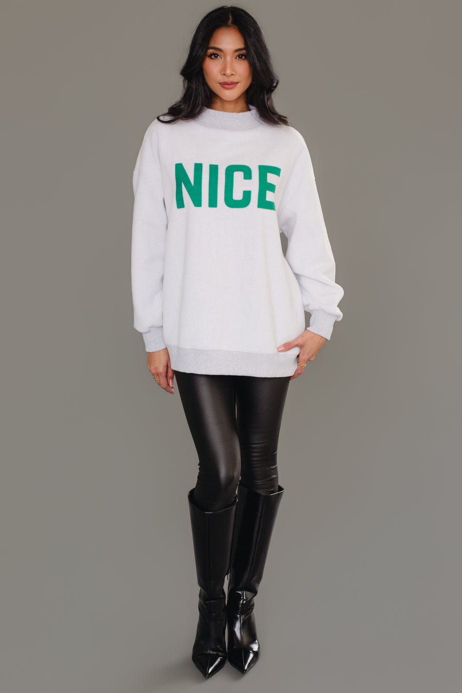 A person wearing a Gray Nice & Naughty Reversible Sweatshirt with the word NICE in large green letters, paired with black leggings and knee-high boots.