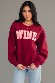 A woman with long dark hair models the Panache Apparel Collection Burgundy Wine Sweatshirt with chenille "WINE" lettering and light blue jeans, standing against a plain gray background.