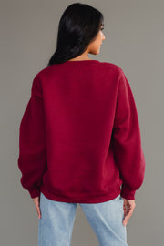 A person with long dark hair models the Panache Apparel Collection Burgundy Wine Sweatshirt, featuring chenille lettering, paired with light blue jeans while facing away from the camera against a plain gray background.