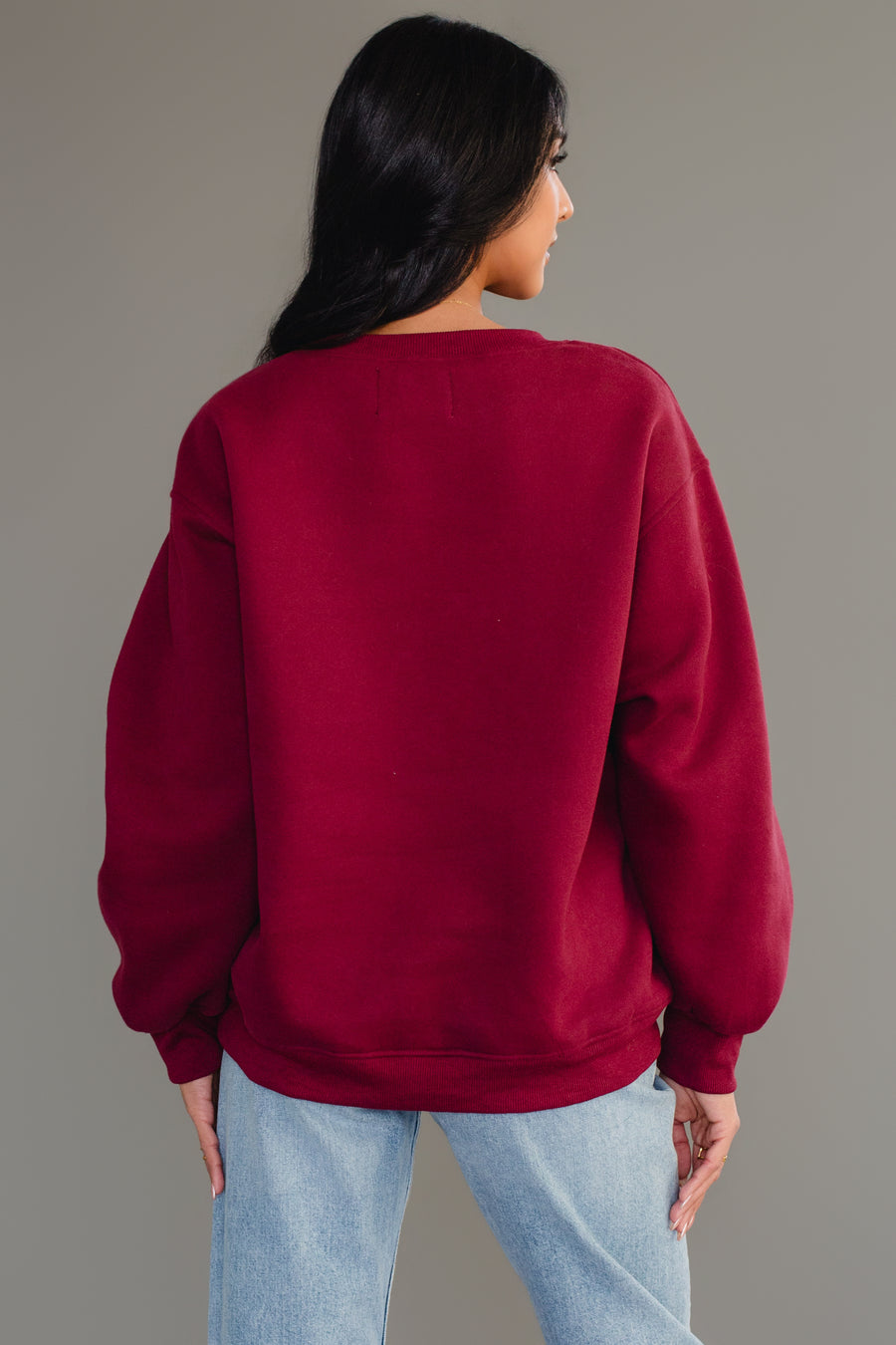 A person with long dark hair models the Panache Apparel Collection Burgundy Wine Sweatshirt, featuring chenille lettering, paired with light blue jeans while facing away from the camera against a plain gray background.