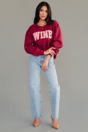 A woman with long dark hair poses against a gray background in the Panache Apparel Collection Burgundy Wine Sweatshirt with chenille WINE lettering, light blue jeans, and nude heels, one hand on her hip.