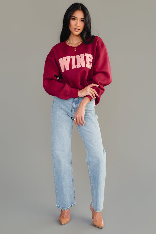 A woman with long dark hair poses against a gray background in the Panache Apparel Collection Burgundy Wine Sweatshirt with chenille WINE lettering, light blue jeans, and nude heels, one hand on her hip.